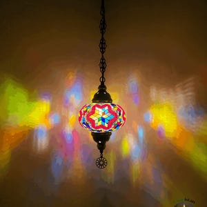 Op de afbeelding: Een kleurrijke moza&iuml;ek glazen hanglamp met een stervormig ontwerp hangt aan een ketting. De lamp werpt kleurrijke reflecties op de muur.