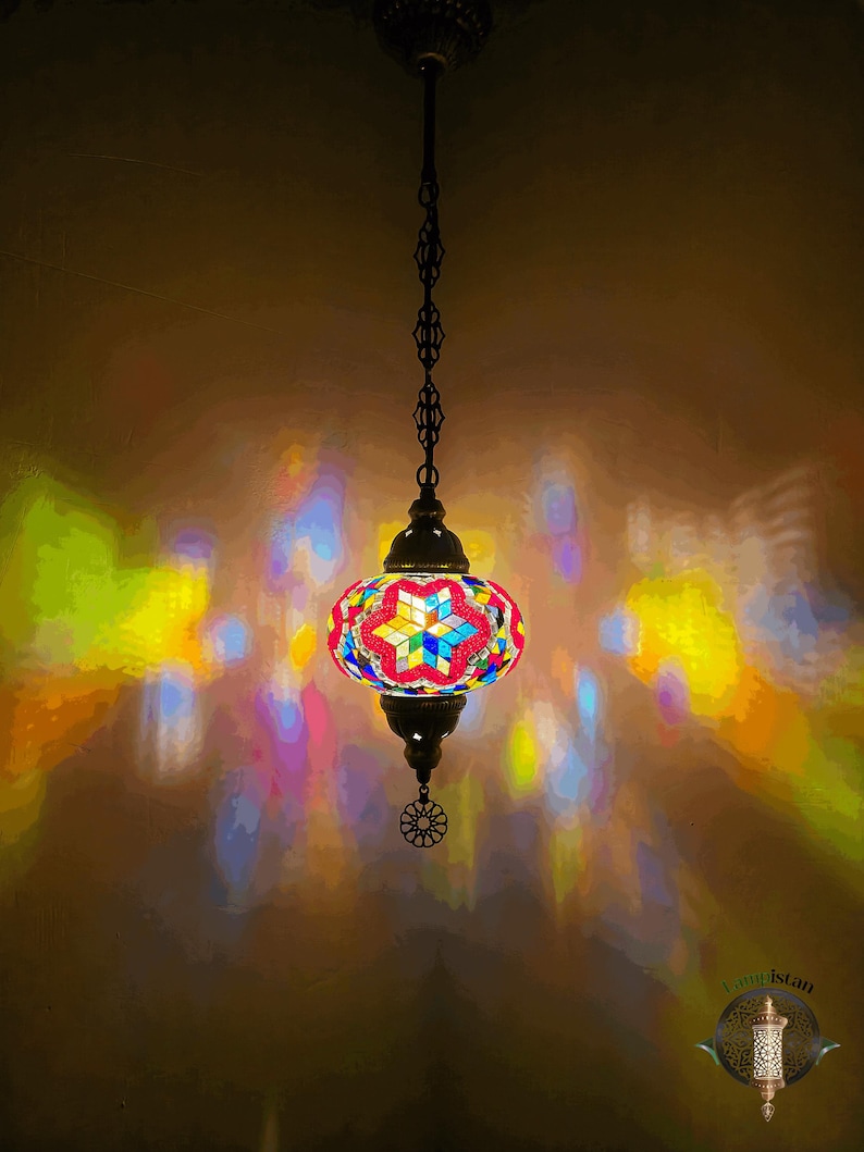 Op de afbeelding: Een kleurrijke moza&iuml;ek glazen hanglamp met een stervormig ontwerp hangt aan een ketting. De lamp werpt kleurrijke reflecties op de muur.