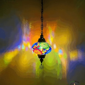 Op de afbeelding: Een kleurrijke moza&iuml;ek glazen hanglamp hangt aan een ketting. De lamp is rond en heeft verschillende kleuren, waaronder blauw, groen, rood en geel. De lamp werpt een kleurrijke gloed op de muur erachter.