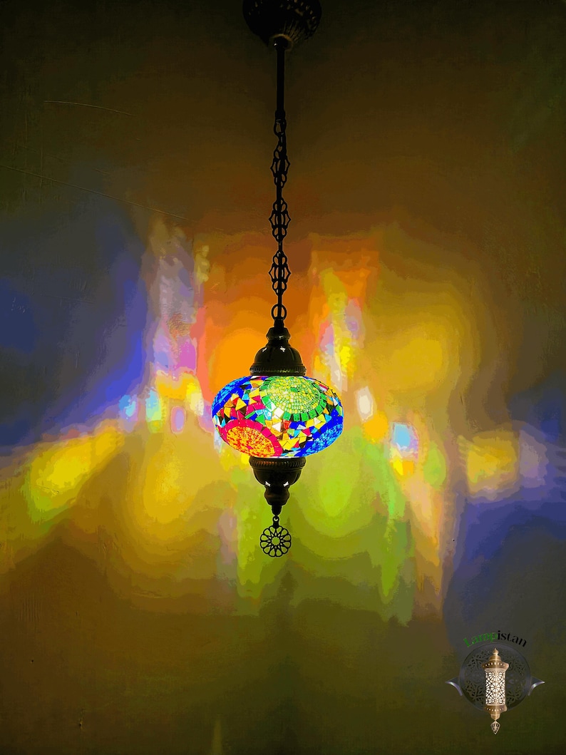 Op de afbeelding: Een kleurrijke moza&iuml;ek glazen hanglamp hangt aan een ketting. De lamp is rond en heeft verschillende kleuren, waaronder blauw, groen, rood en geel. De lamp werpt een kleurrijke gloed op de muur erachter.