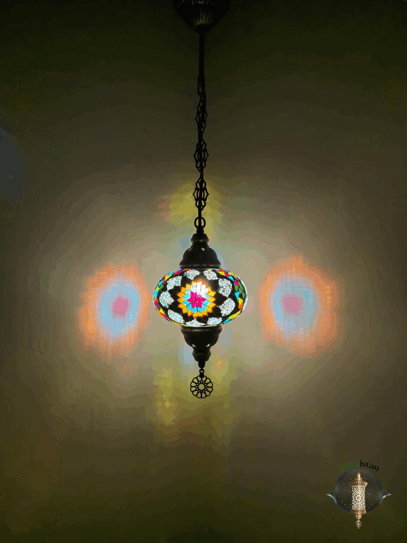 Op de afbeelding: Een kleurrijke moza&iuml;ek glazen hanglamp hangt aan een ketting. De lamp is rond en heeft een geometrisch patroon. Het licht is aan en geeft een warm gloed.