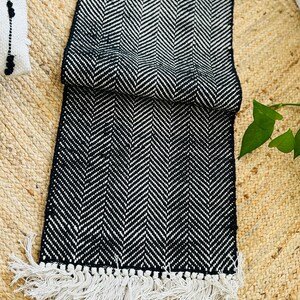 May include: Black and white chevron patterned table runner with white fringe. The runner is folded in half and lying on a woven surface.