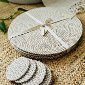 May include: Set of four round woven placemats in a beige and white colour with a white ribbon tied around them. The placemats are on a woven jute surface.