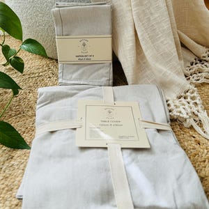May include: A set of grey linen napkins and a grey linen table cover with a brown label that reads "NAPKIN SET OF 6 45cm x 45cm" and "TABLE COVER 150cm x 230cm".