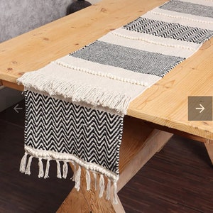 May include: Black and white woven table runner with a chevron pattern and fringe. The runner is on a wooden table.
