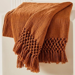 May include: An orange woven throw blanket with a textured weave and a decorative fringe. The fringe has a knotted pattern along the bottom edge. This cosy blanket is a warm, earthy tone, perfect for adding texture to a living space.