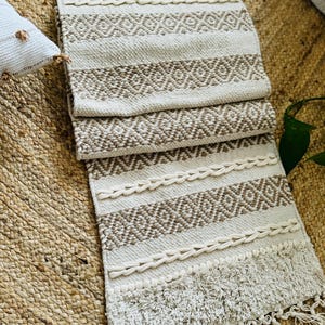 May include: A white and brown woven runner rug with a geometric pattern and fringe. The rug has a textured surface and is made of natural fibres.