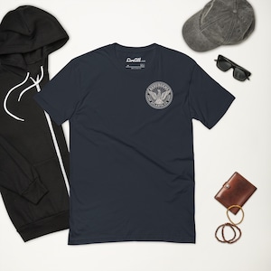 May include: Navy blue t-shirt with a circular emblem featuring an eagle design. The shirt is part of a casual clothing ensemble, alongside a black hoodie, a gray cap, sunglasses, a brown wallet, and bracelets.