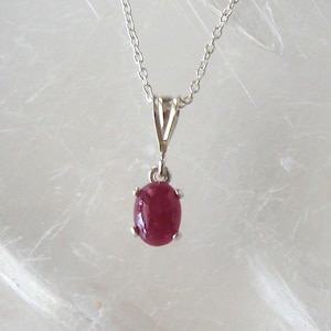May include: A silver necklace featuring a pendant with a vibrant, oval-shaped ruby gemstone. The ruby is a deep red color and is set in a silver bezel. The chain is delicate and hangs from a V-shaped bail.