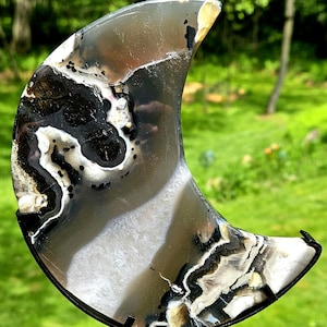 May include: A crescent moon-shaped slice of polished agate stone featuring translucent gray, white, and deep black banding with crystalline inclusions. The stone is mounted on a black metal stand with a circular base and a curved support arm.