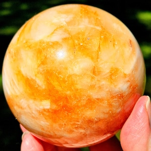 May include: A polished spherical stone featuring a vibrant blend of translucent orange, golden yellow, and creamy white hues. The surface displays natural internal fractures, crystalline textures, and swirling patterns throughout the round mineral form.