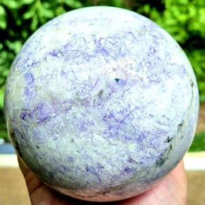 May include: A polished spherical stone featuring a marbled pattern of white and light purple hues. The surface displays intricate dark veining and crystalline textures throughout the round form, creating a natural, multi-toned appearance.