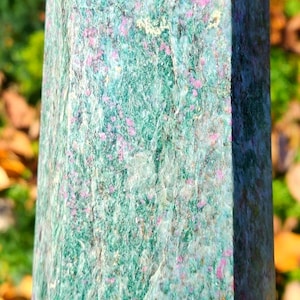 Puede incluir: Un obelisco de cristal alto y facetado. La piedra es una mezcla de tonos verdes, azules y rosas, con una superficie texturizada. El obelisco se sostiene en una mano, con un fondo borroso de follaje.