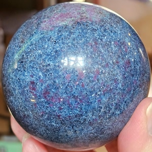 Puede incluir: Una esfera pulida con un patrón moteado de azul profundo y rojo rubí. La bola de piedra preciosa se sostiene en una mano, mostrando su superficie lisa y reflectante. Los colores de la esfera crean un contraste llamativo, ideal para decoración.