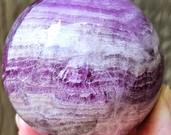 May include: A polished, spherical gemstone with a layered appearance. The sphere features bands of purple and white, creating a marbled effect. The gemstone is held in a hand, showcasing its size and texture.