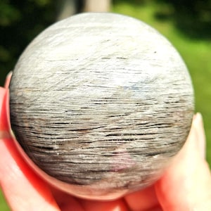 May include: A close-up of a spherical, polished stone. The sphere has a layered, textured appearance with shades of gray and brown. The stone is held in a person's hand, with a blurred green background.