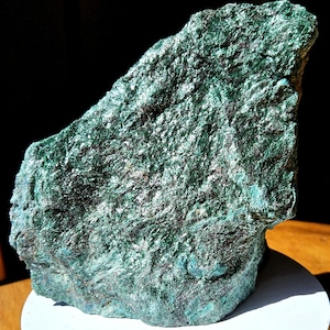 May include: A rough-hewn, irregularly shaped rock formation with a predominantly teal and dark green color palette. The surface has a textured, crystalline appearance, with areas of lighter and darker shades. The rock is displayed against a dark background.