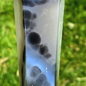 May include: A polished, multi-faceted crystal tower featuring a smooth, tapered point. The stone displays a complex pattern of opaque white, translucent light blue, and deep charcoal black bands, with distinct circular inclusions throughout.