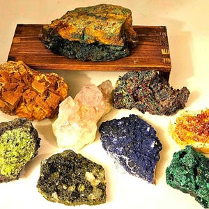 May include: A collection of various colorful mineral specimens, including orange, green, blue, and purple hues. Some are rough and textured, while others have crystalline structures. A wooden display shelf holds a large, layered mineral.