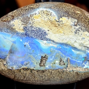 May include: A polished opal stone with a striking display of color. The stone features a vibrant blue and turquoise center, with swirling patterns and iridescent flashes. The outer edges are a mix of brown and gold.