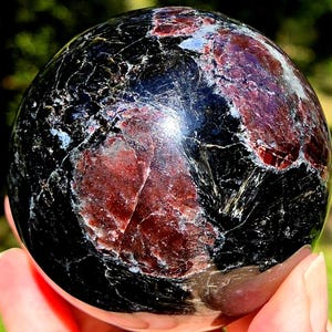 May include: A polished spherical stone featuring a deep black base color with prominent, irregular patches of dark red and maroon mineral inclusions. The surface displays a glossy, reflective texture with visible natural fissures and veins.