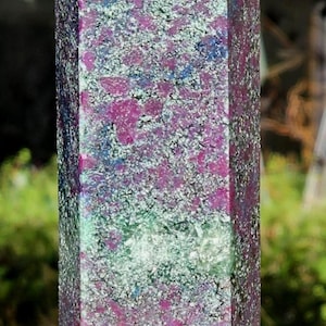 Puede incluir: Un obelisco de cristal rectangular con una superficie pulida. La piedra presenta una mezcla de colores, incluyendo morado oscuro, verde y blanco, con motas rojas. El cristal se sostiene en una mano.