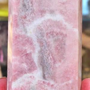 May include: A pink crystal obelisk with a polished, rectangular shape and a pointed top. The stone features a mix of light and dark pink hues with white veining throughout. A natural, decorative item.