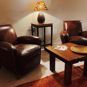 May include: Two brown leather armchairs with tufted backs and a wooden coffee table with a gold tray on top. A lamp with a colourful patterned shade sits on a black metal side table.