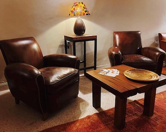 May include: Two brown leather armchairs with tufted backs and a wooden coffee table with a gold tray on top. A lamp with a colorful patterned shade sits on a black metal side table.