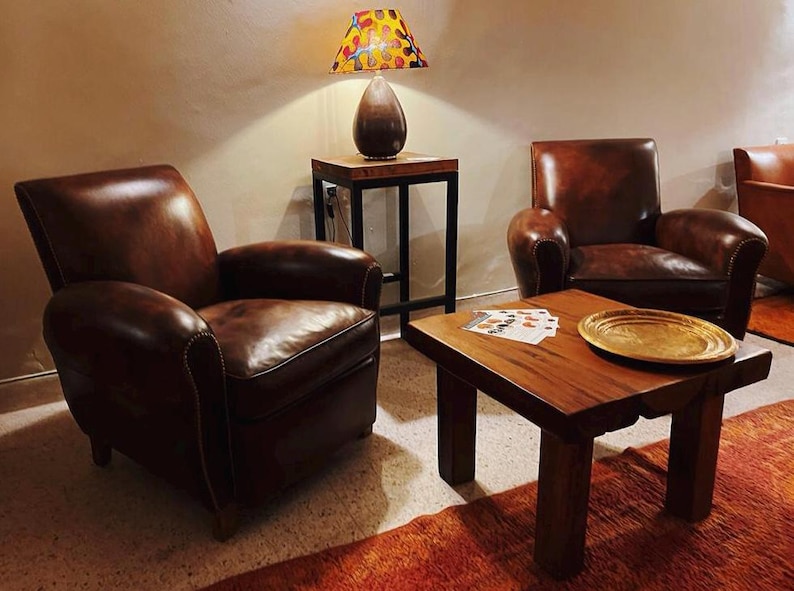 May include: Two brown leather armchairs with tufted backs and a wooden coffee table with a gold tray on top. A lamp with a colourful patterned shade sits on a black metal side table.