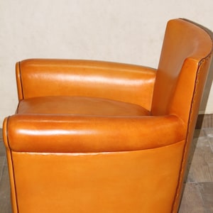 Pu&ograve; includere: Una poltrona in pelle color cuoio con schienale e braccioli arrotondati. La poltrona ha una base in legno marrone scuro ed &egrave; rivestita in pelle liscia e morbida.