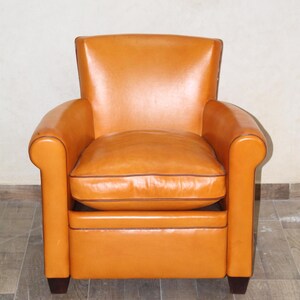 Pu&ograve; includere: Una poltrona in pelle color cuoio con un cuscino del sedile trapuntato e gambe in legno marrone scuro.