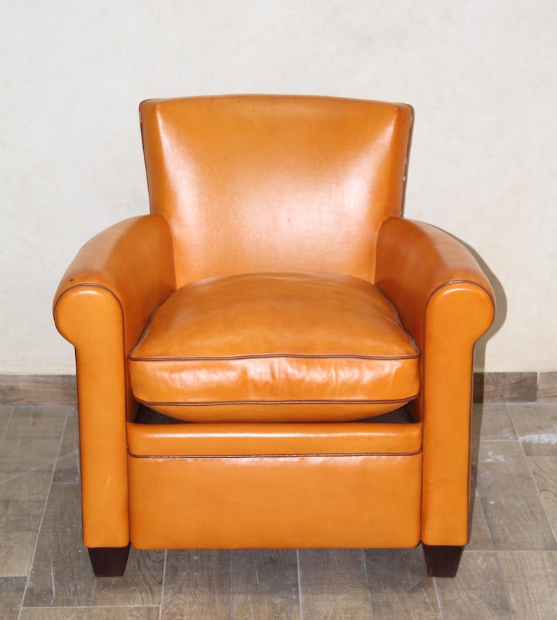 Pu&ograve; includere: Una poltrona in pelle color cuoio con un cuscino del sedile trapuntato e gambe in legno marrone scuro.