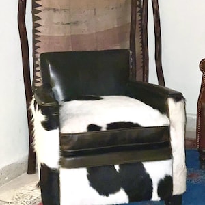 Può includere: Una poltrona in pelle nera con un motivo in pelle di mucca bianca e nera. La poltrona ha una struttura in legno e si trova su un tappeto.