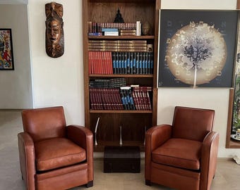 May include: Two brown leather armchairs with wooden legs, a wooden bookshelf filled with books, a wooden sculpture of a person's face, and a framed print of a tree with a globe design.