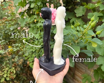 May include: A black candle shaped like a person labeled male and a white candle shaped like a person labeled female, both standing on a heart-shaped base. The candles feature wicks at the top and are positioned back-to-back on the dark base.
