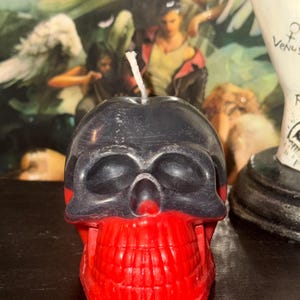 May include: A black and red skull-shaped candle with a white wick. The skull has a black top half and a red bottom half with a detailed tooth design.