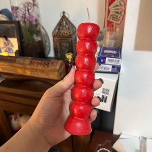 May include: A vibrant red candle with a unique, stacked design. The candle is cylindrical, featuring a series of rounded segments, and has a wick at the top. The candle is held in a hand, with other objects in the background.