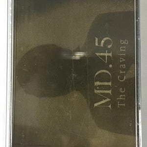 May include: A vintage cassette tape in a clear plastic case. The tape label is dark with the text "MD.45" stacked above "The Craving" in a serif font. The case is rectangular.