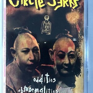 May include: A cassette tape case with the band name "Circle Jerks" in yellow, stylized lettering. The album cover features two bald figures with a dark background and the words "oddities, abnormalities, and curiosities."