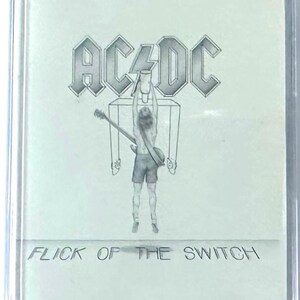 May include: A cassette tape with the album art for AC/DC's "Flick of the Switch." The cover features the band's logo above a drawing of a person holding a guitar. The album title is printed at the bottom.