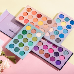 May include: A collection of colorful eyeshadow palettes. Each palette features a variety of shades, including matte and shimmer finishes. The palettes are arranged on a pink and yellow surface. The text "COLOR BOX BEAUTY GLAZED" is visible on one of the palettes.