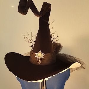 May include: A brown velvet witch hat with a wide brim and a tall pointed crown. The hat is decorated with a black tulle veil, a twig and leaf embellishment, and a silver and gold moon and star pendant.