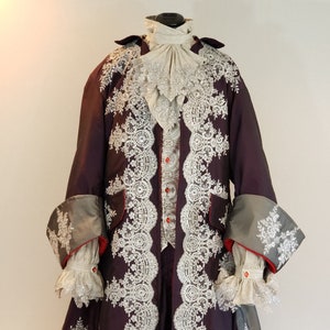 Puede incluir: Una chaqueta de brocado morado y plateado con ribetes de encaje y detalles rojos. La chaqueta tiene un cuello alto y mangas largas con puños. La chaqueta se lleva sobre una camisa blanca con una corbata de encaje.