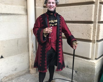 May include: A man in a red and black brocade coat with a white wig and a cane. He is holding a glass of champagne.