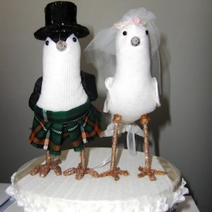 May include: A wedding cake topper featuring a bride and groom dressed as birds. The bride is wearing a white veil and a flower crown. The groom is wearing a black top hat and a green kilt. The cake topper is made of fabric and is standing on a white cake.