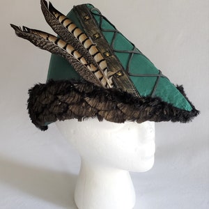May include: A green felt hat with a black faux fur brim and gold accents. Two brown and white feathers are attached to the top of the hat.