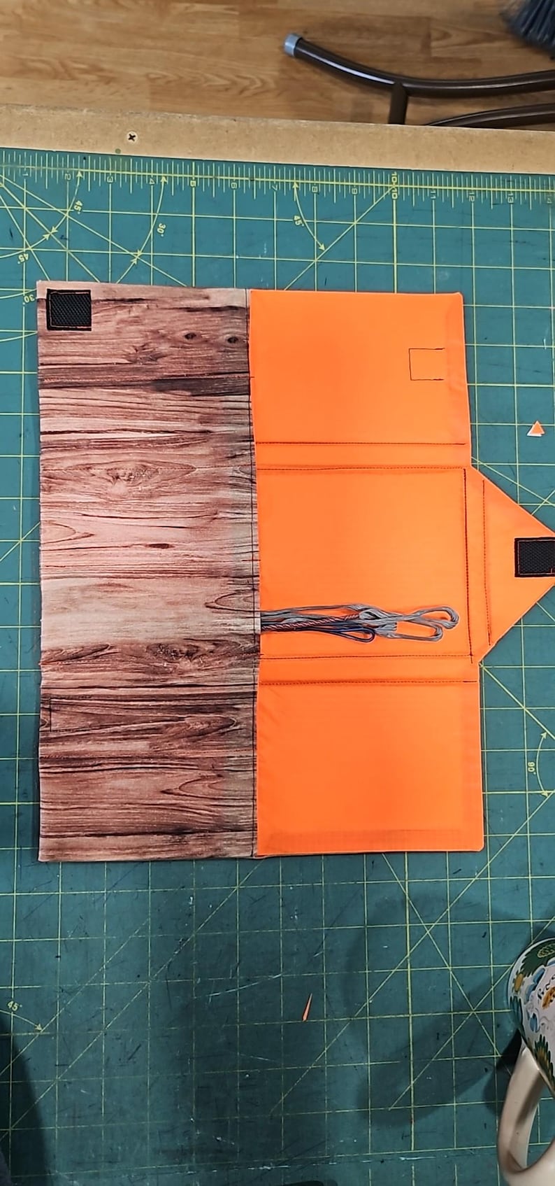 May include: A fabric organiser, half wood-effect and half bright orange. The organiser is open, showing pockets and a bundle of grey and blue threads. A black square is on the wood-effect side. The organiser is on a green surface.