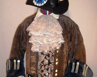 May include: A brown velvet jacket with gold trim and buttons. The jacket has a white lace cravat and a white lace ruffle at the cuffs. The jacket is worn over a brown and gold patterned vest. The person is wearing a black hat with a blue and white ribbon.