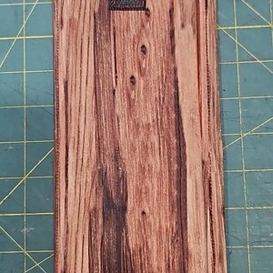 May include: A rectangular phone case with a wood grain pattern. The top is orange with a black square. The case is approximately 25 cm long.